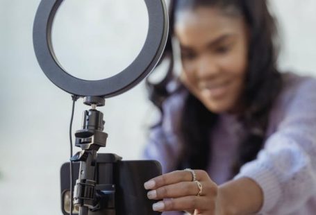 Social Media Mobile - Cheerful young African American female blogger in stylish sweater smiling while setting up camera of smartphone attached to tripod with ring light before recording vlog