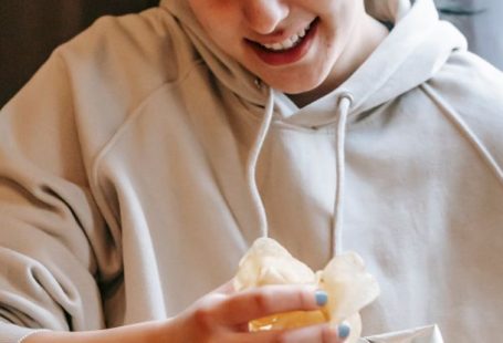 Content Repurpose - Smiling plump female wearing comfy hoodie eating crispy yummy potato chips while sitting in light room