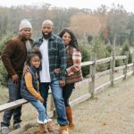 Evergreen Content - Young content African American parents embracing daughter and son near wooden fence on tree farm while looking at camera