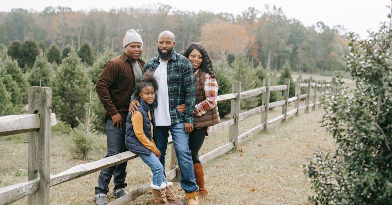 Evergreen Content - Young content African American parents embracing daughter and son near wooden fence on tree farm while looking at camera